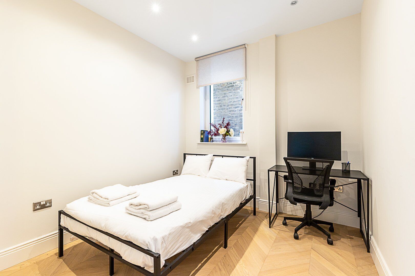 Un dormitorio con cama, escritorio y silla, ventana con vista a un muro de ladrillos, y una mesita con un florero.