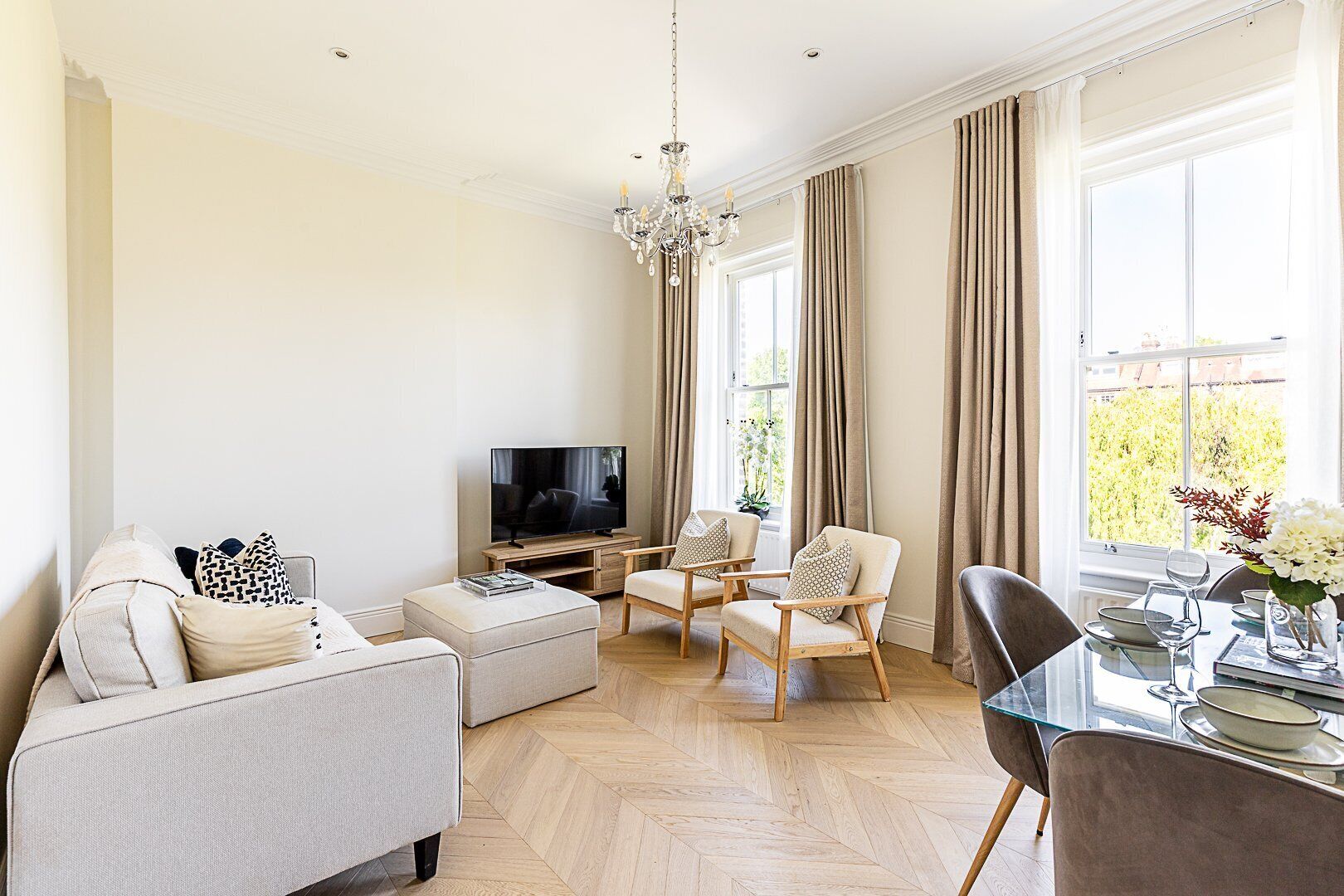 A modern living room with a flat-screen TV, a sofa, two armchairs, a glass dining table, and a chandelier.