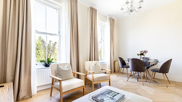A modern living room with two armchairs, a dining table with chairs, a chandelier, and large windows with curtains. - Bright & Modern Hampstead Apartment (London)