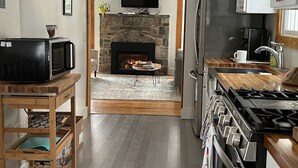 A modern kitchen with a stone fireplace, a television mounted above it, and a wooden cart with kitchen items. - Private Oasis Awaits ! Hot Tub, Fire Pit, nap in the Hammock under the trees (Canadensis)