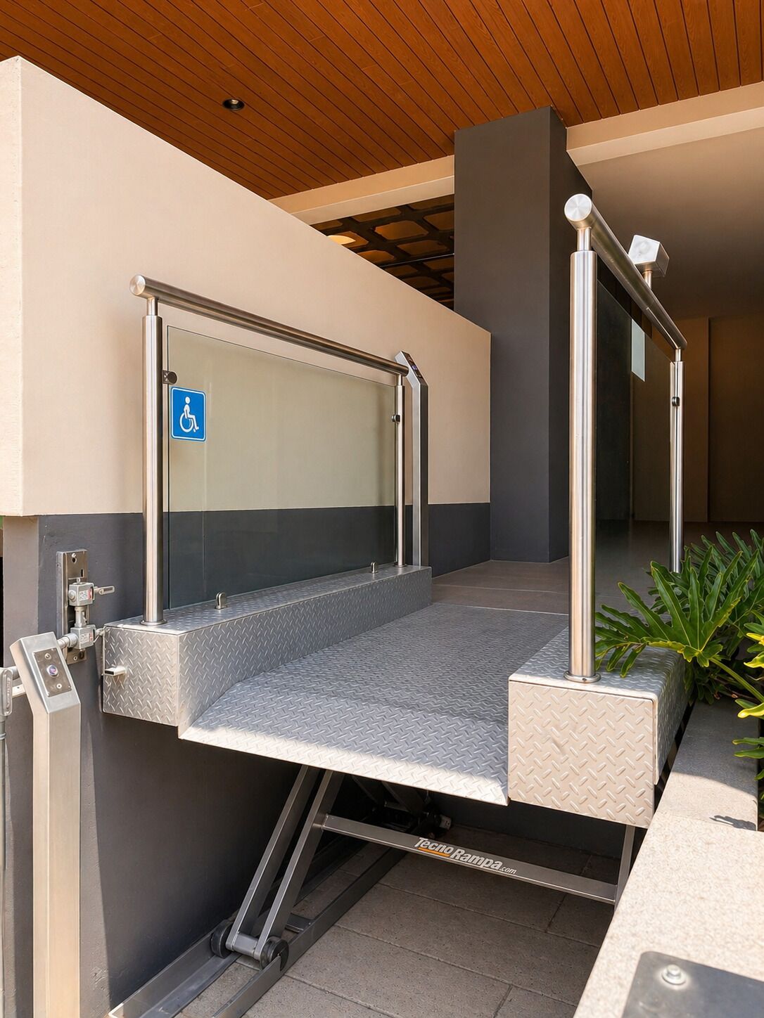 A modern outdoor elevator with a wheelchair-accessible ramp, metal handrails, and a control panel.