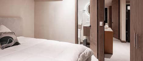 A modern bedroom with a bed, a bedside table, a mirror, and a bathroom visible through an open door.