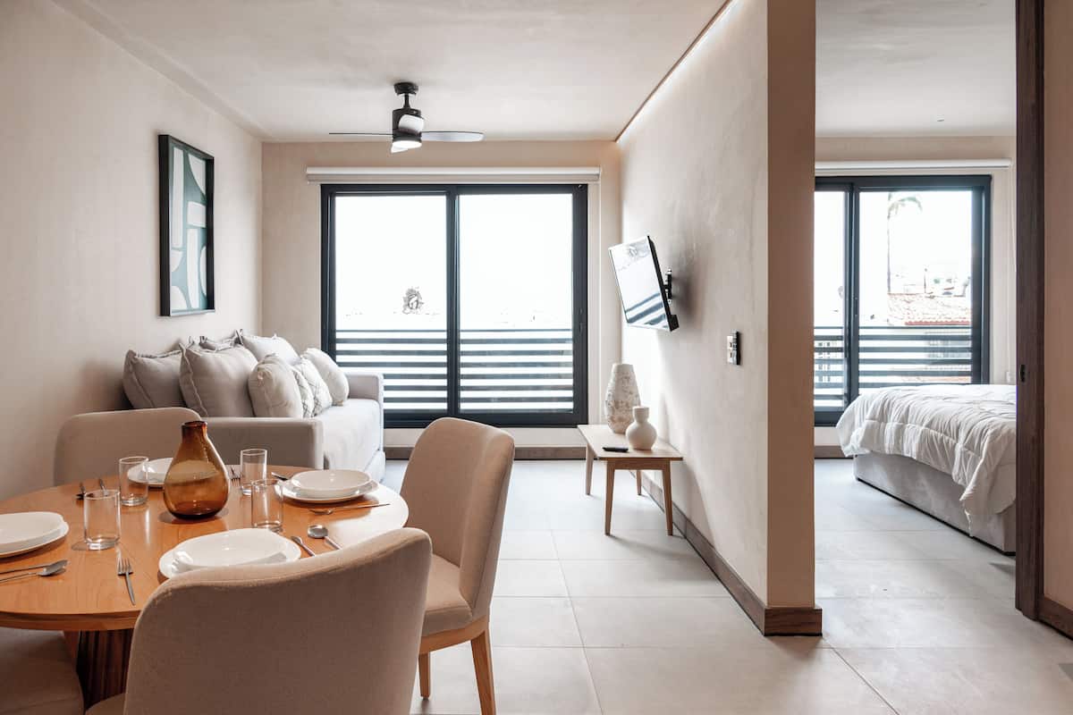 Una habitación de hotel moderna con zona de comedor, sofá, cama y ventanas amplias.