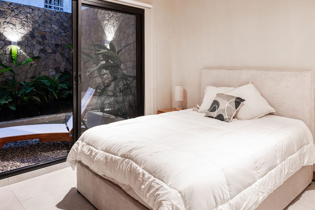 Un dormitorio con una cama grande, una mesita de noche, una lámpara y vistas a una pared de piedra y plantas.