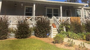 A two-story house with a covered porch, a front yard with a lawn, and a small garden area. - !!POOL!! PEACEFUL FARMHOUSE IN SHALLOTTE! 
5 MINS TO BEACHES AND OUR SWEET TOWN! (Shallotte)