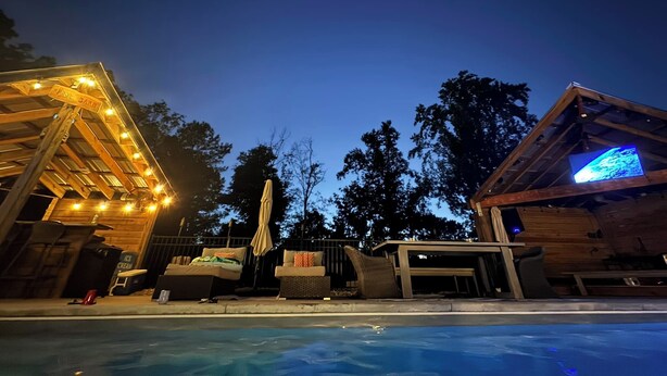 A poolside area with outdoor seating, a wooden structure with a sign, and a building with a digital display. - !!POOL!! PEACEFUL FARMHOUSE IN SHALLOTTE! 
5 MINS TO BEACHES AND OUR SWEET TOWN! (Shallotte)