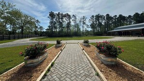 A paved pathway leading to a building with a covered entrance, surrounded by a grassy area and trees. - !!POOL!! PEACEFUL FARMHOUSE IN SHALLOTTE! 
5 MINS TO BEACHES AND OUR SWEET TOWN! (Shallotte)