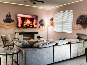 Una sala de estar con un sofá seccional, un televisor montado en la pared, un ventilador de techo y una ventana con persianas.