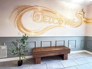 Un área de recepción con un banco, un mural en la pared con la palabra 'WELCOME' y una planta en maceta.