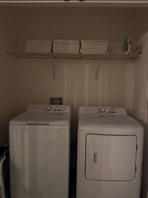 Two front-loading washing machines with control panels, a shelf above holding folded towels, and a power outlet on the wall. - 9BR Fairytale Kingdom Sleeps 24 Pool Near Disney (Kissimmee)