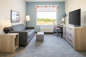 A compact living space with a sofa, a desk, a television, and a window with a view of greenery. - Home2 Suites by Hilton Lee's Summit Kansas City (Lee's Summit)