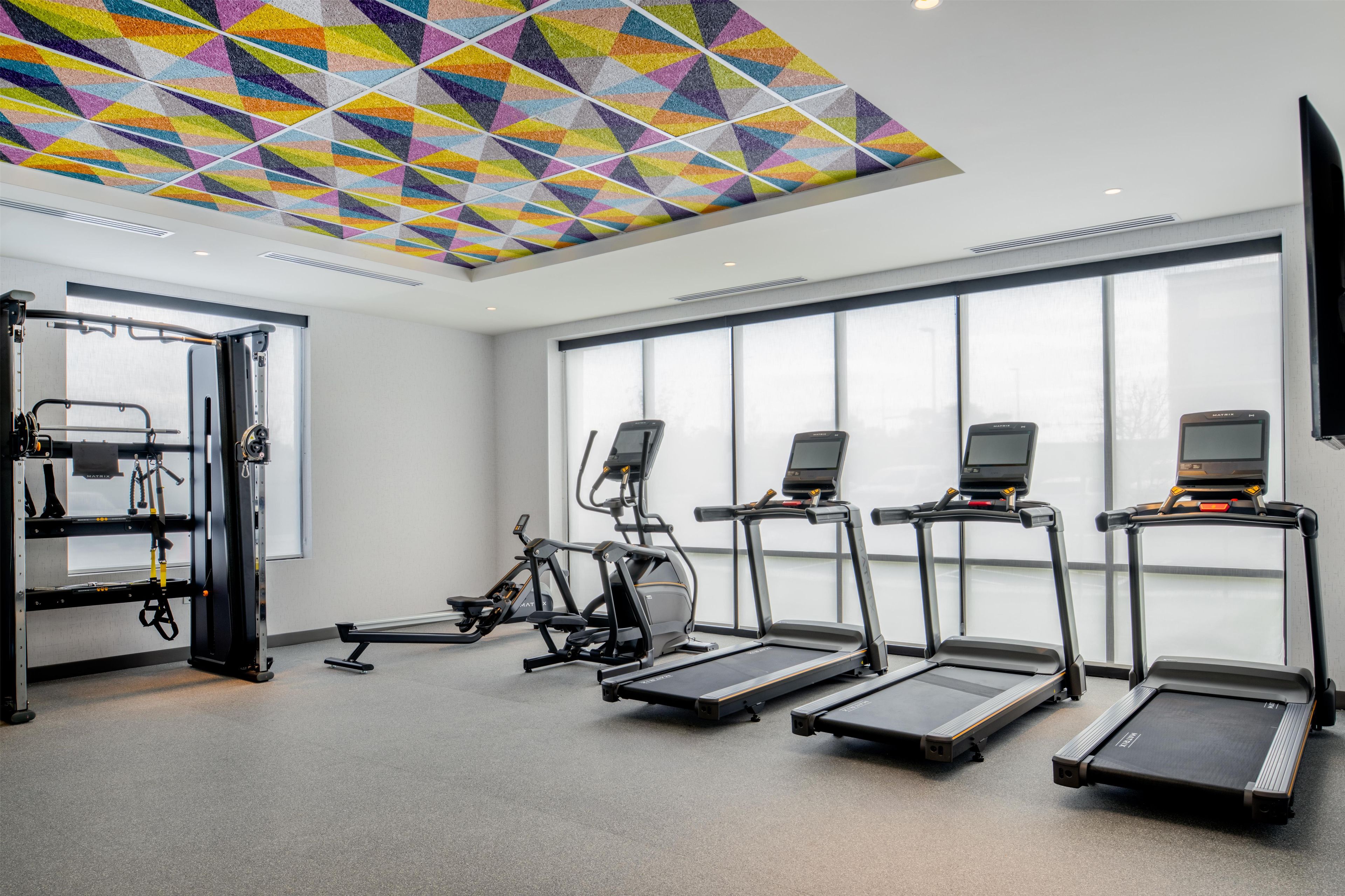 Un gimnasio con cintas de correr, bicicletas estáticas y un techo con un patrón geométrico colorido.