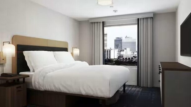 A hotel room with a bed, nightstands, a desk, a chair, a window with a city view, and a flat-screen TV. - Homewood Suites by Hilton Oakland Airport Alameda (Oakland)
