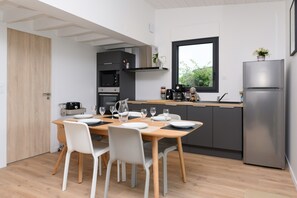 Une cuisine moderne avec une table à manger pour quatre personnes, un réfrigérateur et une fenêtre donnant sur un espace verdoyant.