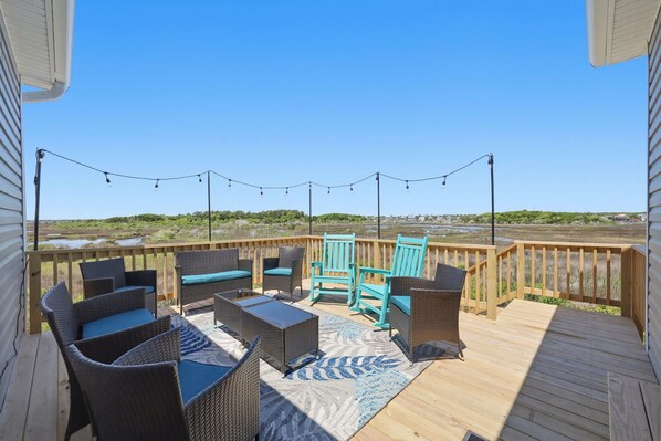 A deck with wicker furniture, a patterned rug, and string lights. - Turtle LIVIN | ICW Views, Sleeps 11, Games & Relax (North Topsail Beach)