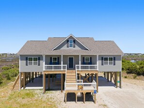 A two-story beach house with a front porch, wooden deck, and a view of the beach.