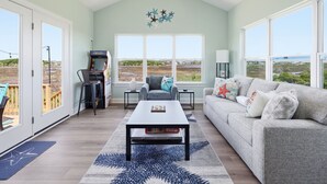 A spacious living room with a large window, a ceiling fan, a gaming console, and a sofa with star-themed pillows. - Turtle LIVIN | ICW Views, Sleeps 11, Games & Relax (North Topsail Beach)