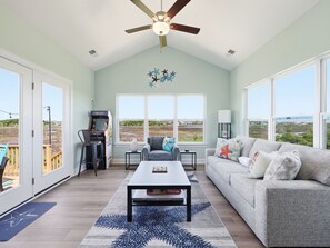 A spacious living room with a large window, a ceiling fan, a gaming console, and a sofa with star-themed pillows.