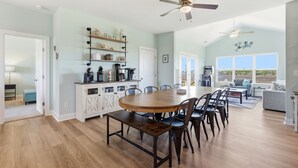 A spacious dining area with a wooden table and chairs, a kitchen with white cabinetry, and a view through large windows. - Turtle LIVIN | ICW Views, Sleeps 11, Games & Relax (North Topsail Beach)