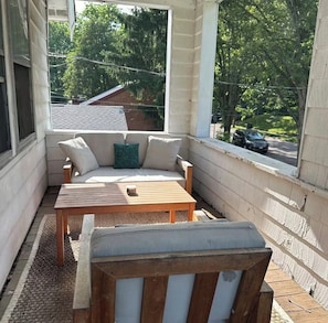 Un espace extérieur aménagé, abrité, avec une table en bois et un canapé, une fenêtre donnant sur la rue, et une vue sur des arbres et un bâtiment au loin.