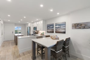 A modern kitchen with white cabinets, a central island, and a dining area with wicker chairs. - Luxury Waterfront 2BR/2BA Marco Island condo (Marco Island)