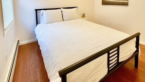 A bedroom with a bed, a nightstand, a painting on the wall, and a window with blinds.