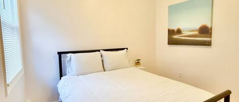 A bedroom with a bed, a nightstand, a painting on the wall, and a window with blinds.