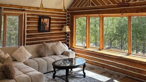 A log cabin interior with a large window, a chandelier, a sofa, and a table. - 🌲Chalet Spa & Sauna Getaway in Luarentide– Sainte-Lucie 🌲 Val-David, Val-Morin (Sainte-Lucie-des-Laurentides)