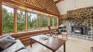 A log cabin interior with a stone fireplace, wooden beams, large windows, and a cozy seating area. - 🌲Chalet Spa & Sauna Getaway in Luarentide– Sainte-Lucie 🌲 Val-David, Val-Morin (Sainte-Lucie-des-Laurentides)