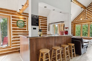 Ein Holzblockhaus-Innenraum mit einer Holztheke, Barhocker, einer Couch und einem großen Fenster.