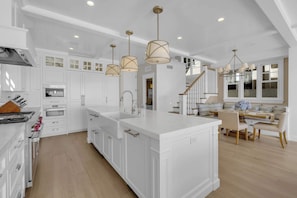 A modern kitchen with white cabinetry, a large island, pendant lighting, and a dining area with a wooden table and chairs. - Luxury Newport Beach House with Rooftop Sunset Deck, Hot Tub and Game Room (Newport Beach)