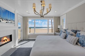A bedroom with a large bed, a fireplace, a chandelier, a view of the beach, and a window with curtains. - Luxury Newport Beach House with Rooftop Sunset Deck, Hot Tub and Game Room (Newport Beach)