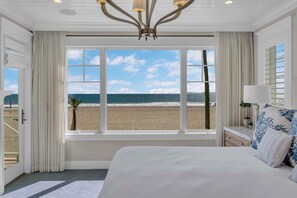 A bedroom with a large window overlooking a beach, a bed with white bedding, and a chandelier. - Luxury Newport Beach House with Rooftop Sunset Deck, Hot Tub and Game Room (Newport Beach)