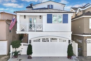 A two-story house with a white exterior, a garage, and a small balcony. - Luxury Newport Beach House with Rooftop Sunset Deck, Hot Tub and Game Room (Newport Beach)