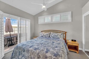 Une chambre à coucher comprenant un lit, un ventilateur de plafond, une table de chevet, une table avec une chaise et une fenêtre avec des stores.