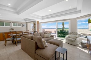A spacious living room with a large sectional sofa, a dining area with wooden chairs, and large windows offering a view of the beach. - Beachfront Oasis: Rooftop and Firepit, Sleep 12 (Newport Beach)