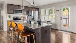 Una cocina moderna con una isla central, taburetes de bar, electrodomésticos de acero inoxidable y vistas a través de una puerta.