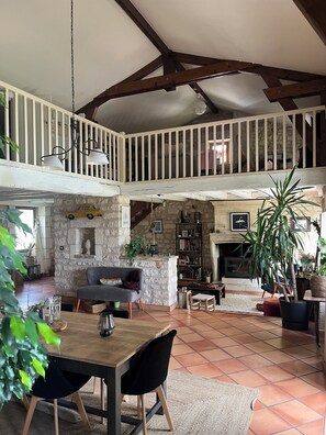 Un'ampia zona giorno con soffitto alto, travi in legno a vista, camino in pietra e un balcone con ringhiera.