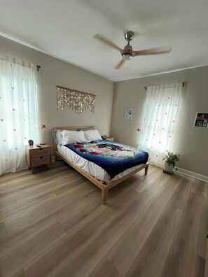 A bedroom with a wooden bed, a ceiling fan, a bedside table, a nightstand, and a potted plant. - Cozy Anime Beach House - Quick Walk to Beach (Bolivar Peninsula)