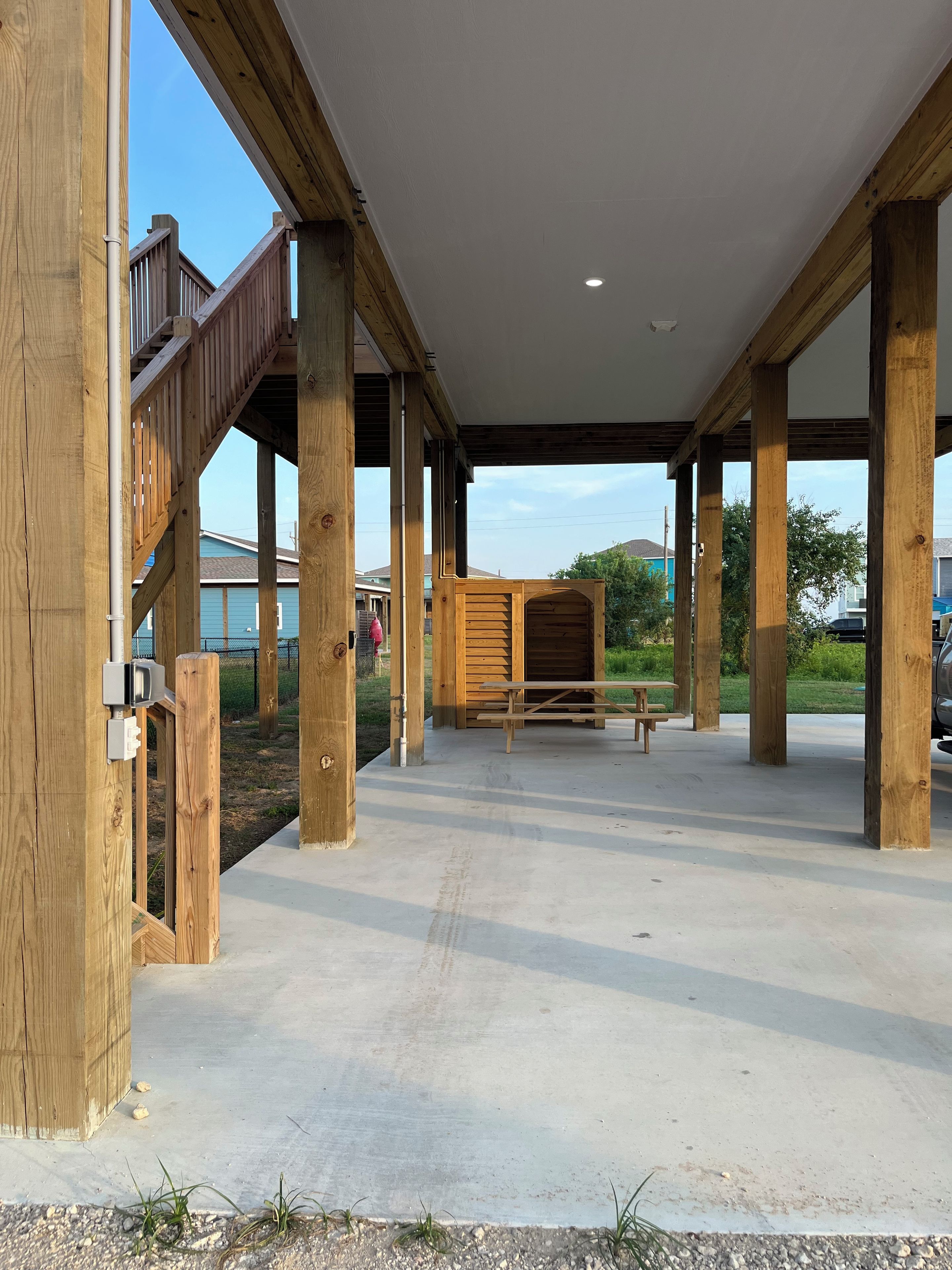 Un espacio al aire libre techado con vigas de madera y piso de concreto, que incluye una mesa de picnic y una estructura de madera al fondo.