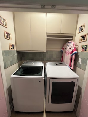 A laundry area with two white washers and dryers, a shelf above, and a pink jersey hanging on the wall. - Cozy Anime Beach House - Quick Walk to Beach (Bolivar Peninsula)