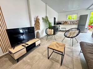 Ein modernes Wohnzimmer mit einem Flachbildfernseher, einem Holztisch und zwei schwarzen Stuhlgestellen. Im Hintergrund ist eine Küche mit einer Waschmaschine und einem Kühlschrank zu sehen.