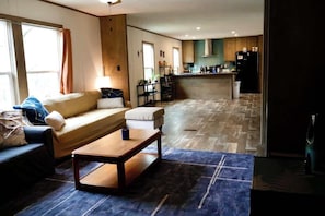 A living room with a sofa, a coffee table, and a rug. There is a kitchen area with cabinets and appliances in the background.
