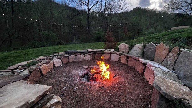 Un foc circular de pedra amb flames, envoltat d'una paret de pedra, situat en una zona verda amb arbres al fons.