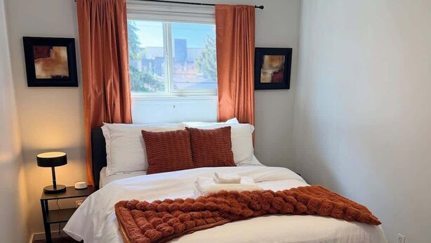 A bedroom with a bed, a window with curtains, a bedside table with a lamp, and two wall-mounted picture frames. - Cozy 2BR Near Zoo, Downtown & Max Bell (Calgary)