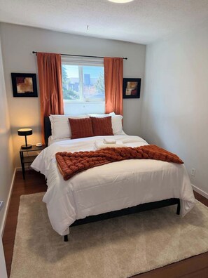 A bedroom with a bed, a window with curtains, a bedside table with a lamp, and two wall-mounted picture frames.