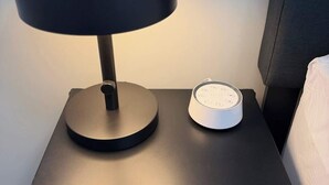 A bedside table with a lamp and a white device, possibly a speaker or alarm clock, on a black nightstand. - Cozy 2BR Near Zoo, Downtown & Max Bell (Calgary)