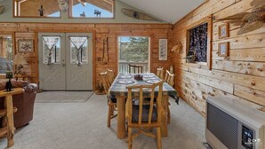 Interior de uma cabana de madeira com mesa de jantar, cadeiras, lareira e uma janela grande.