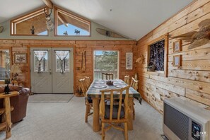 A wooden cabin interior with a dining table, chairs, a fireplace, and a large window. - Wandering Moose Riverfront Hideaway (Soldotna)
