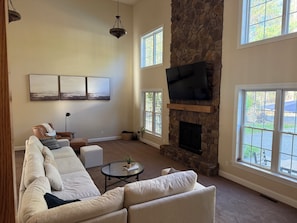 A spacious living room with a stone fireplace, large windows, a flat-screen TV, and a sectional sofa.